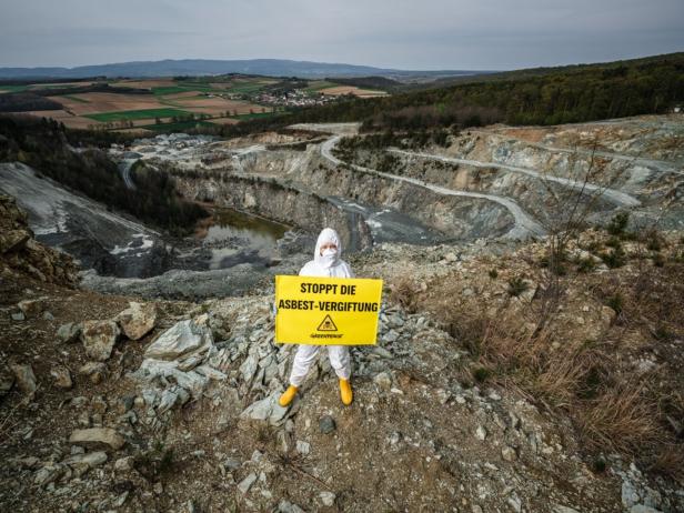 Eine Person in Schutzkleidung steht in einer Steinbruchlandschaft und hält ein gelbes Schild mit der Aufschrift „Stoppt die Asbest-Vergiftung“.