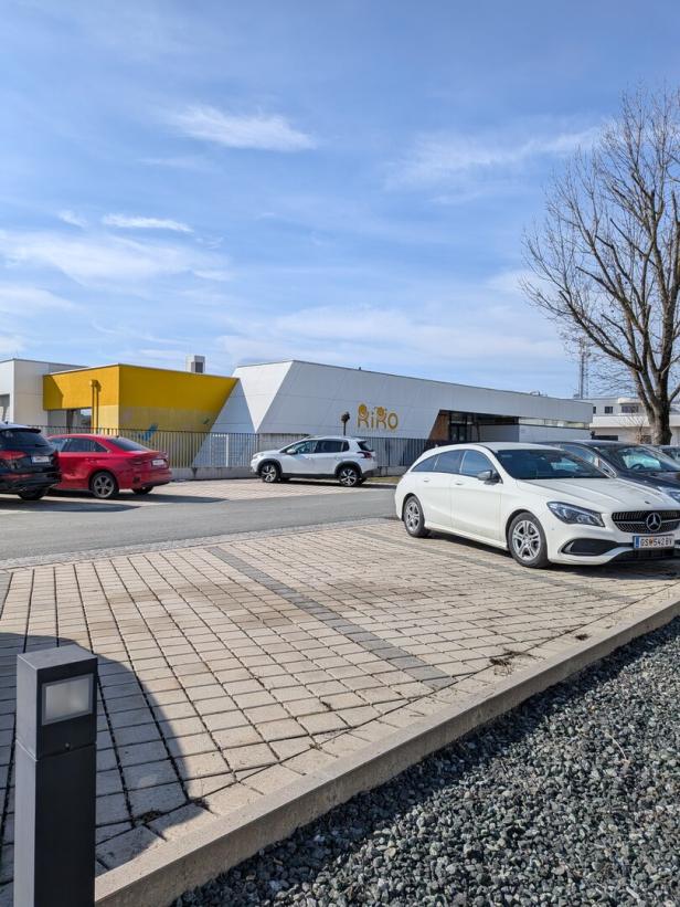 Mehrere Autos parken vor einem modernen Gebäude mit gelber und weißer Fassade, daneben steht ein kahler Baum.