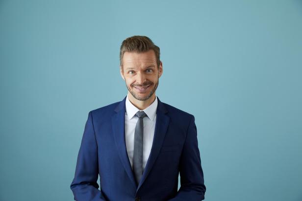 Ein Mann im blauen Anzug mit weißem Hemd und Krawatte lächelt vor hellblauem Hintergrund.