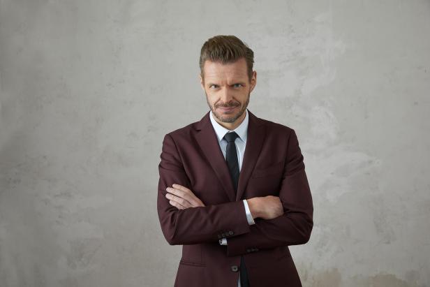 Mann im Anzug mit verschränkten Armen vor grauer Wand.