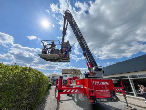Ein Feuerwehrfahrzeug mit Drehleiter hebt zwei Feuerwehrleute in einem Korb in die Höhe bei sonnigem Wetter.