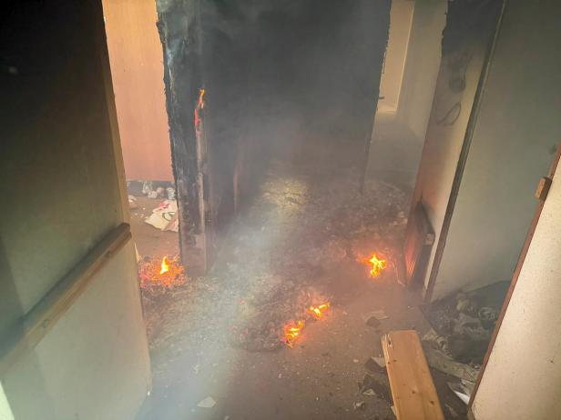 Flammen und Rauch breiten sich in einem stark beschädigten, verrußten Flur eines Gebäudes aus.