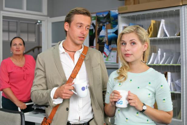 Zwei junge Menschen unterhalten sich mit Kaffeetassen in der Hand, im Hintergrund steht eine Frau in einem Büro.