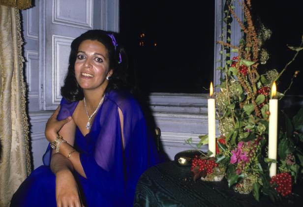 Christina Onassis in einem blauen Kleid mit Schmuck sitzt neben einem Fenster und einem Gesteck mit Kerzen.