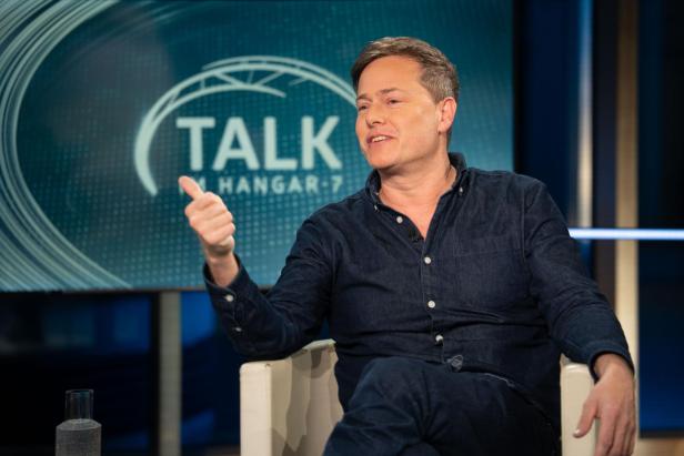Ein Mann diskutiert in einer Talkshow vor dem Bildschirm mit der Aufschrift "Talk im Hangar-7".