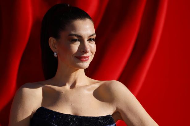 Anne Hathaway lächelt vor einem roten Hintergrund in einem trägerlosen, dunklen Kleid.