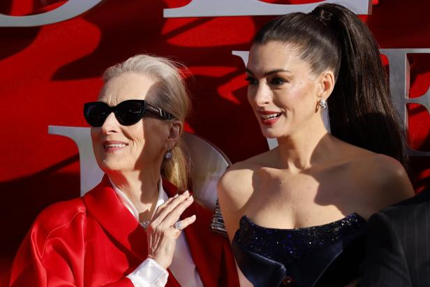 Meryl Streep mit Sonnenbrille und Anne Hathaway lächeln vor rotem Hintergrund.
