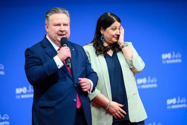 Ein Mann im Anzug spricht ins Mikrofon, während eine Frau neben ihm sich emotional das Gesicht berührt.