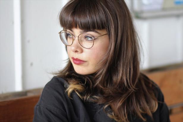 Junge Frau mit braunen Haaren, Brille und Pony schaut nach links und trägt einen dunklen Pullover.