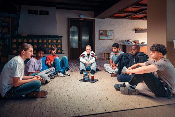 Sieben Personen sitzen im Kreis auf dem Boden, in der Mitte stehen ein gerahmtes Foto und eine Kerze.