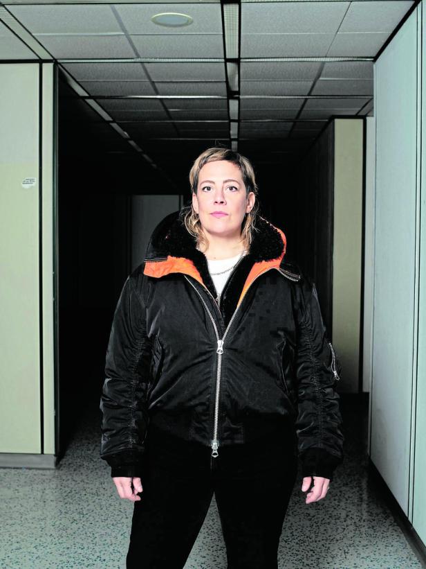 Eine Person mit heller Jacke und ernstem Blick steht in einem Flur mit weißen Wänden und Deckenplatten.