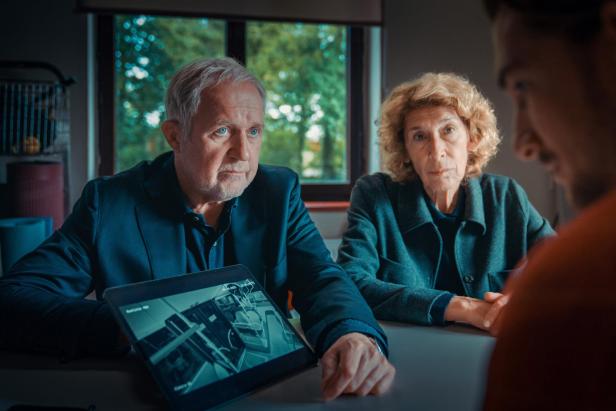 Zwei ältere Personen sitzen ernsthaft an einem Tisch und zeigen einem Mann auf einem Tablet eine Überwachungskameraaufnahme.