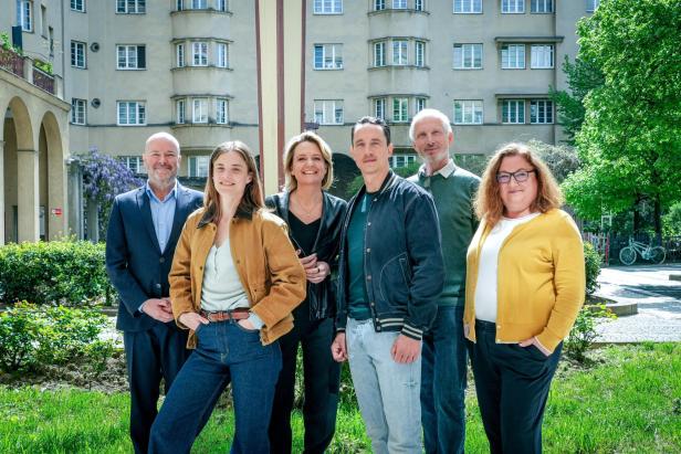Eine Gruppe von sechs Personen steht vor einem Gebäude mit vielen Fenstern.