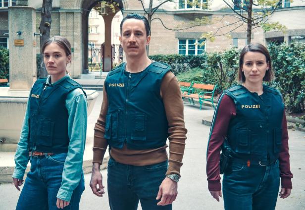 Drei Polizisten mit kugelsicheren Westen stehen ernsthaft in einem Innenhof mit Bänken und Bäumen.