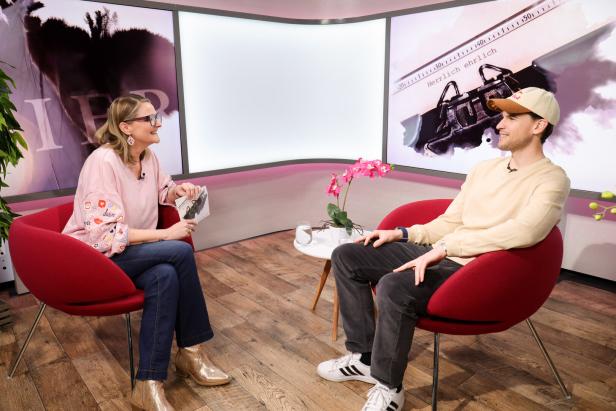 Zwei Personen sitzen sich in roten Sesseln gegenüber und unterhalten sich in einem modern eingerichteten Studio mit Blumen auf dem Tisch.