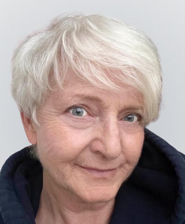 Ältere Frau mit kurzem, weißem Haar und blauen Augen lächelt leicht und trägt eine dunkle Kapuze.