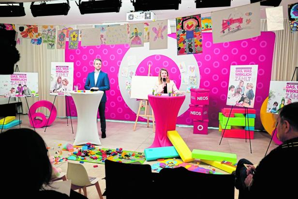 Zwei Personen sprechen vor pinkfarbenem Hintergrund mit Kinderzeichnungen und NEOS-Logos.