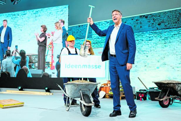 Ein Mann im Anzug hält einen Spaten über einer Schubkarre mit Sand, daneben steht ein Schild mit der Aufschrift „Ideenmasse“.