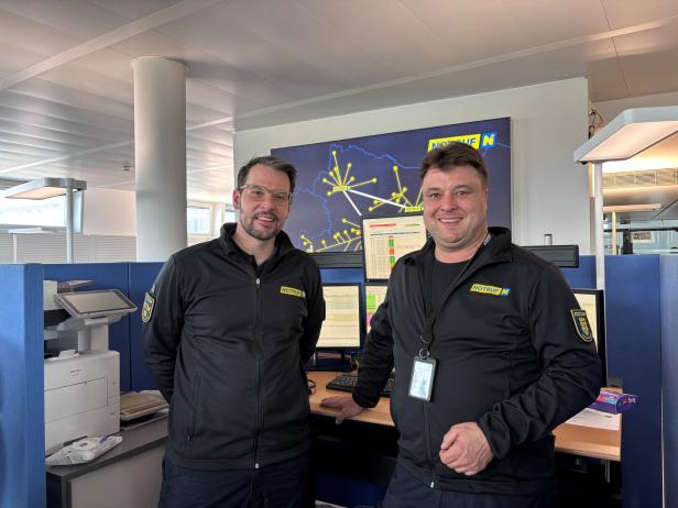 Zwei Männer in schwarzer Einsatzkleidung stehen lächelnd in einer Leitstelle vor Monitoren und einer Karte mit gelben Linien.