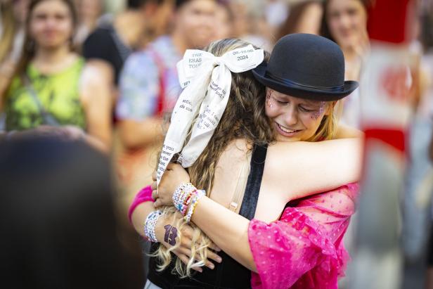 Zwei junge Menschen umarmen sich herzlich, eine trägt einen schwarzen Hut und Glitzer im Gesicht, die andere ein großes Schleifenband im Haar.