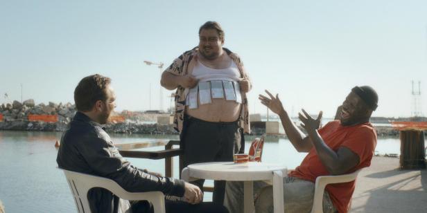 Drei Männer sitzen an einem Tisch am Wasser, einer zeigt lachend mehrere Ausweise unter seinem hochgezogenen Hemd.