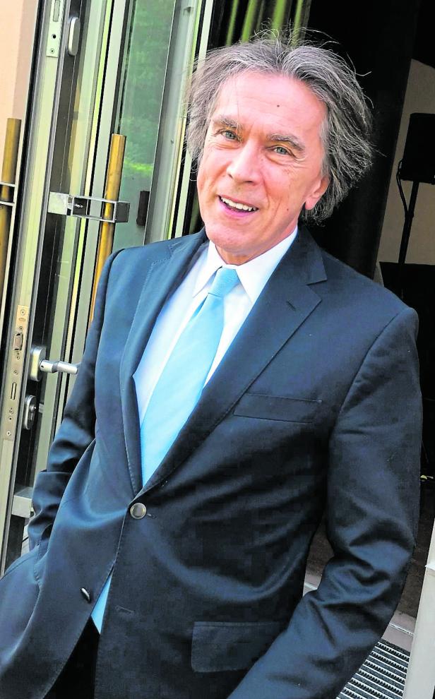 Emil Brix, ein mit grauen Haaren trägt einen dunklen Anzug und eine hellblaue Krawatte und steht lächelnd vor einer offenen Tür.