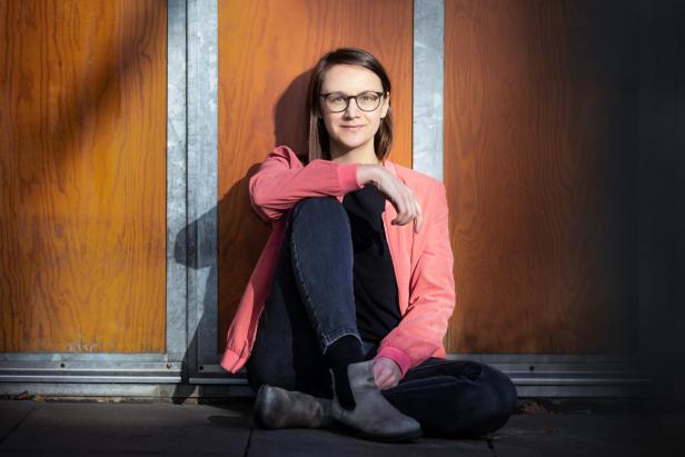 Johanna Sebauer, eine Frau mit Brille und rosa Jacke, sitzt entspannt vor einer orange-braunen Holzwand auf dem Boden.