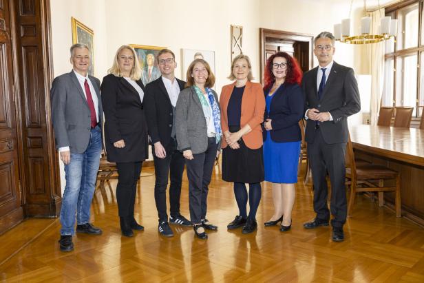 Eine Gruppe von sieben Personen posiert in einem Raum mit Holzboden und Gemälden.
