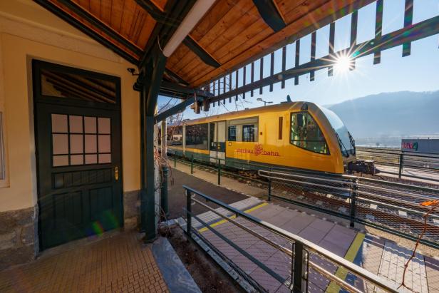 Ein gelber Zug der Wachau Bahn steht an einem sonnigen Bahnsteig mit Blick auf Berge im Hintergrund.