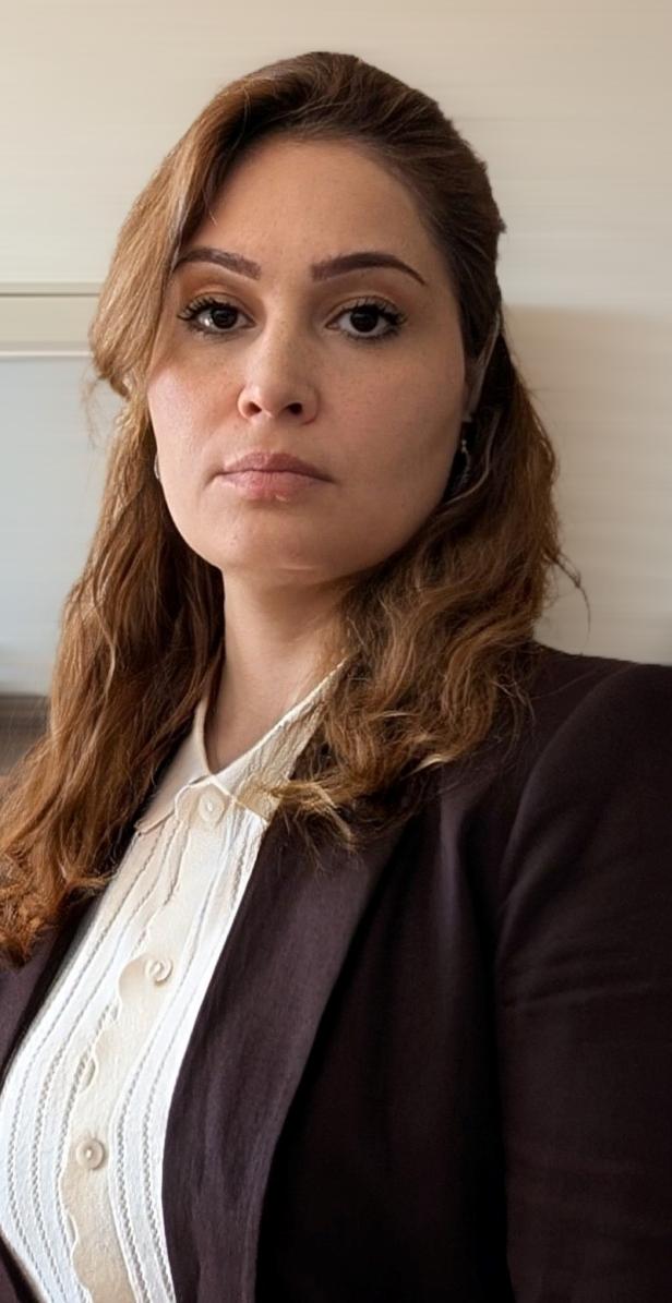 Frau mit langen braunen Haaren trägt eine weiße Bluse und einen dunklen Blazer, schaut ernst nach vorn.