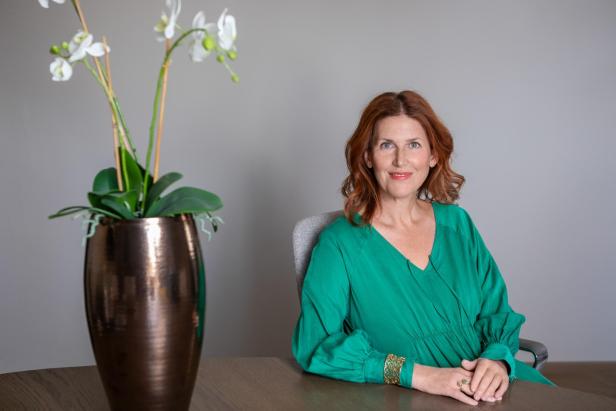 Eine Frau mit roten Haaren sitzt an einem Tisch, trägt ein grünes Kleid und im Vordergrund steht eine Orchidee in einer Vase.