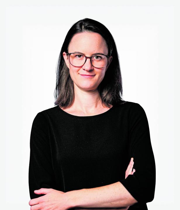 Frau mit schulterlangem dunklem Haar, Brille und schwarzem Oberteil steht mit verschränkten Armen vor hellem Hintergrund.