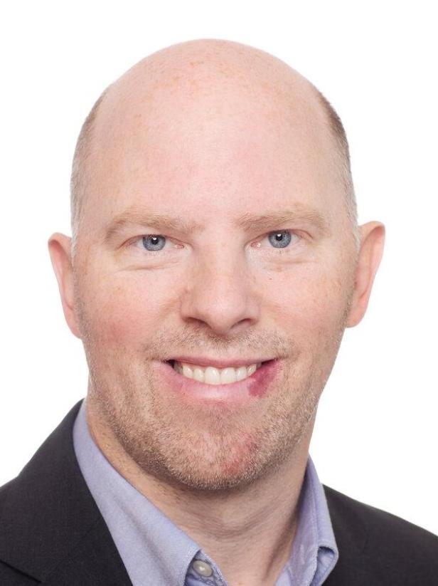 Ein lächelnder Mann mit hellen Augen und Anzug vor weißem Hintergrund.