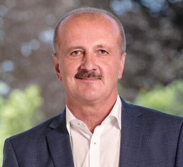 Ein Mann mit Schnurrbart und Anzug steht vor einem unscharfen, grünen Hintergrund.