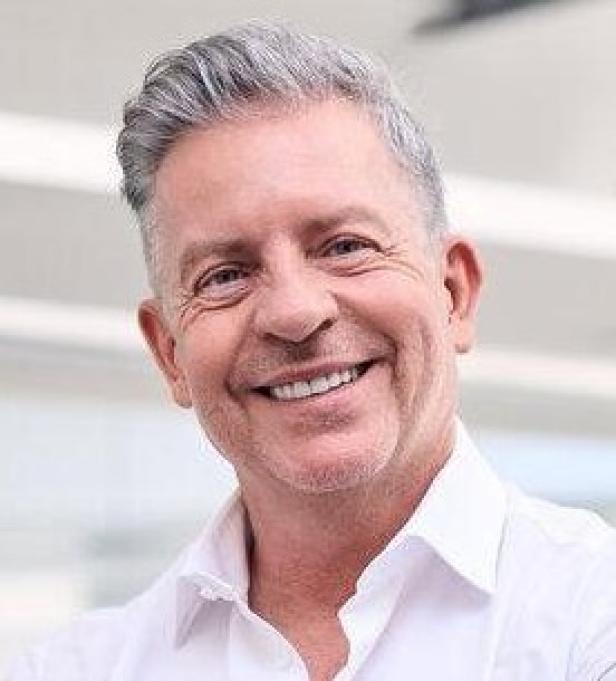 Ein lächelnder Mann mit grauen Haaren und weißem Hemd steht mit verschränkten Armen da.
