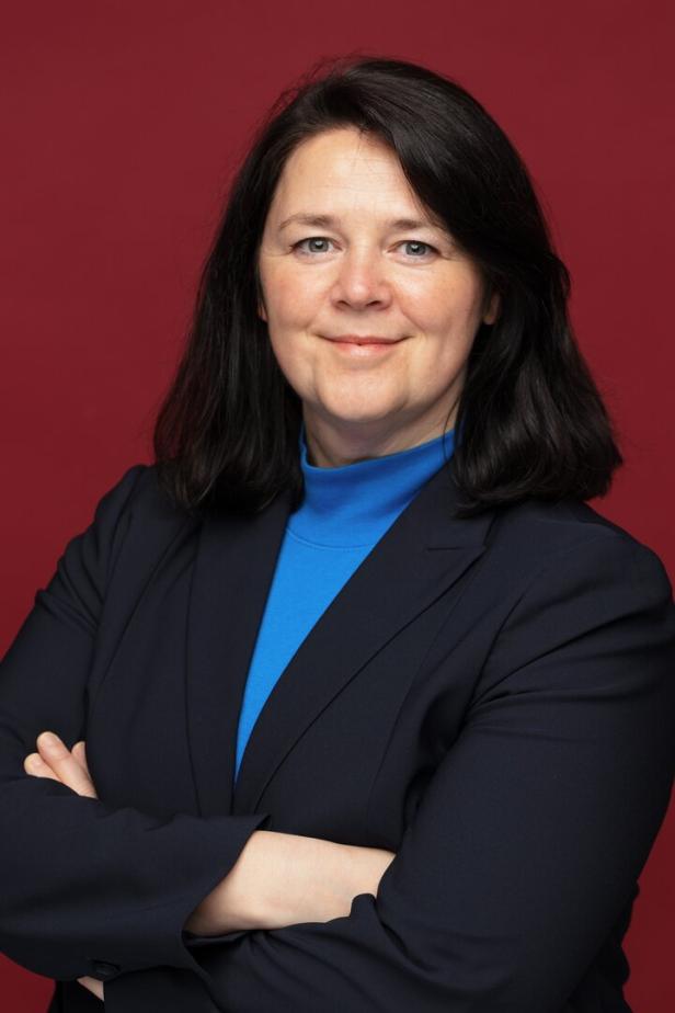 Frau mit dunklen Haaren und blauem Rollkragenpullover unter einem dunklen Blazer steht vor rotem Hintergrund.