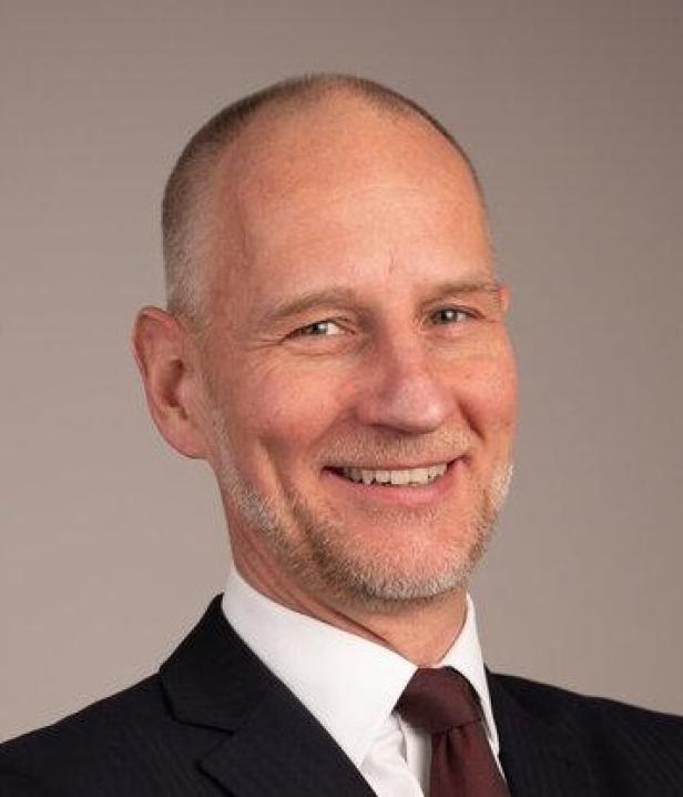 Ein Mann im Anzug mit verschränkten Armen lächelt vor neutralem Hintergrund.