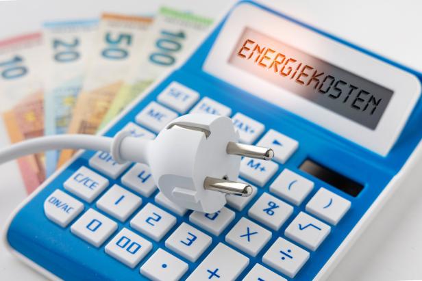 Ein Stecker liegt auf einem blauen Taschenrechner, dessen Display "Energiekosten" anzeigt, im Hintergrund Euroscheine.