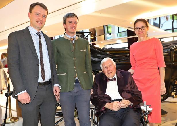 Drei stehende Erwachsene und ein älterer Mann im Rollstuhl posieren festlich gekleidet in einem hellen Innenraum.