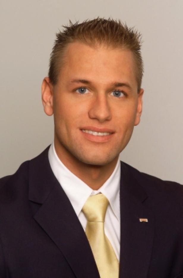 Ein Mann mit blonden Haaren, Anzug und Krawatte lächelt in die Kamera.