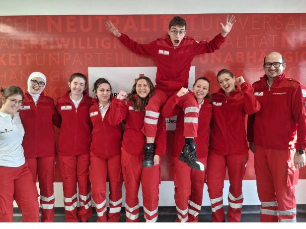 Neun Menschen in roten Rettungsdienst-Uniformen stehen lachend zusammen, eine Person sitzt auf den Schultern einer anderen.