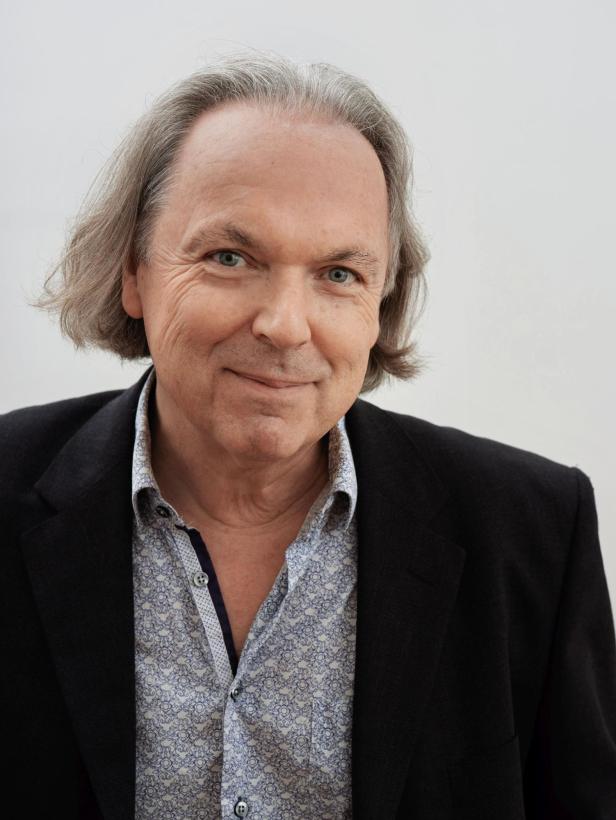 Ein älterer Mann mit grauen, schulterlangen Haaren trägt ein gemustertes Hemd und ein dunkles Sakko, er lächelt leicht.