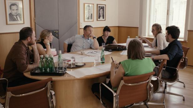 Sieben Personen sitzen an einem Konferenztisch und diskutieren, auf dem Tisch liegen Papiere und Wasserflaschen.