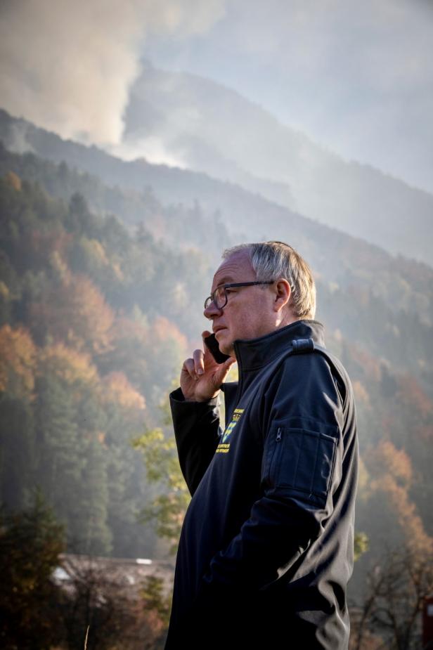Ein Mann mit Brille telefoniert im Freien, im Hintergrund rauchende Berge und herbstlicher Wald.