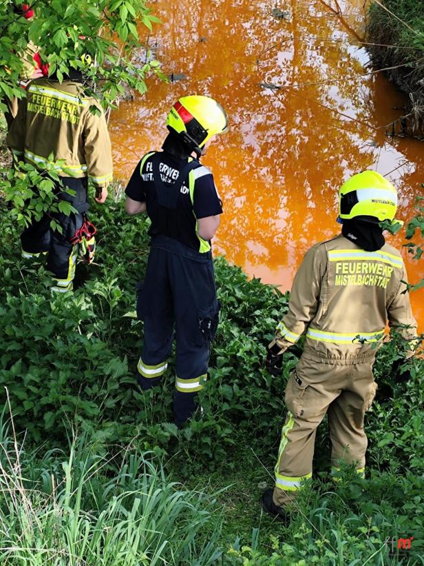 Drei Feuerwehrleute stehen am Ufer eines orange gefärbten Flusses in Mistelbach.