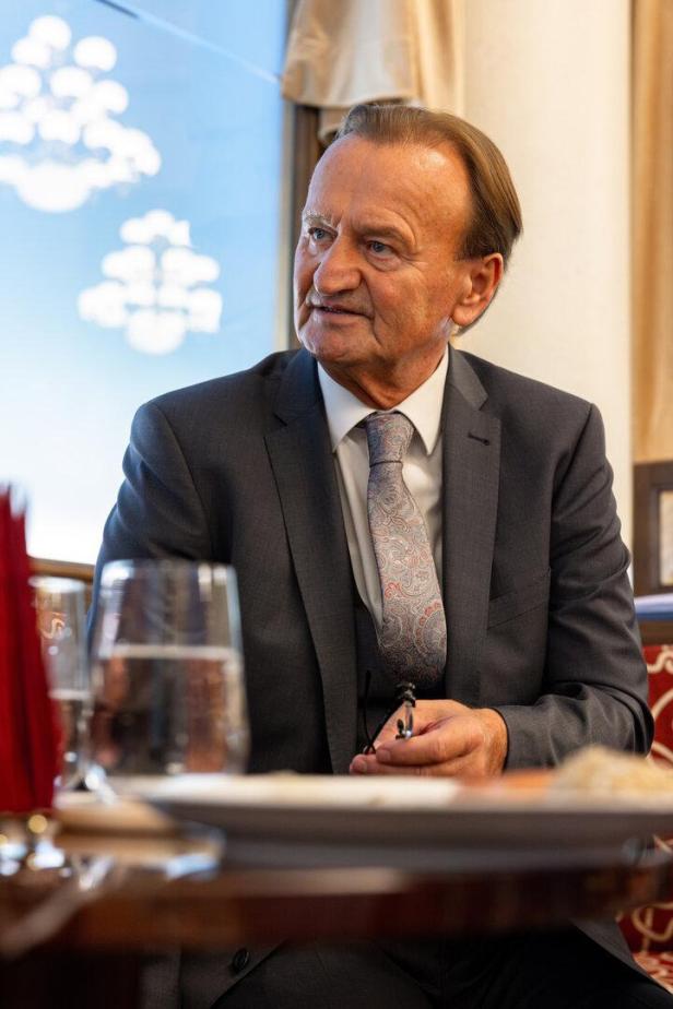 Ein älterer Mann im Anzug sitzt an einem gedeckten Tisch und hält eine Brille in der Hand.