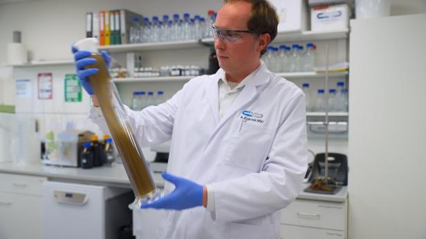 Ein Mann im Laborkittel und mit Schutzbrille hält einen großen Messzylinder mit brauner Flüssigkeit in einem Labor.