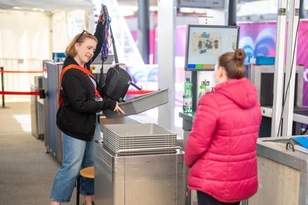 Eine Frau legt ihre Tasche in eine Sicherheitswanne, während eine andere Person in roter Jacke daneben steht.