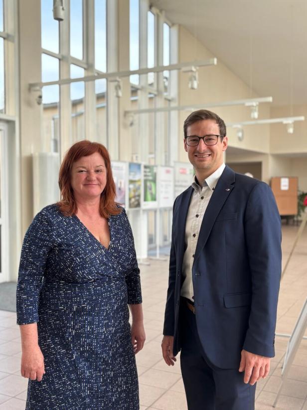 Eine Frau im gemusterten Kleid und ein Mann im Anzug stehen lächelnd in einem hellen, modernen Foyer.