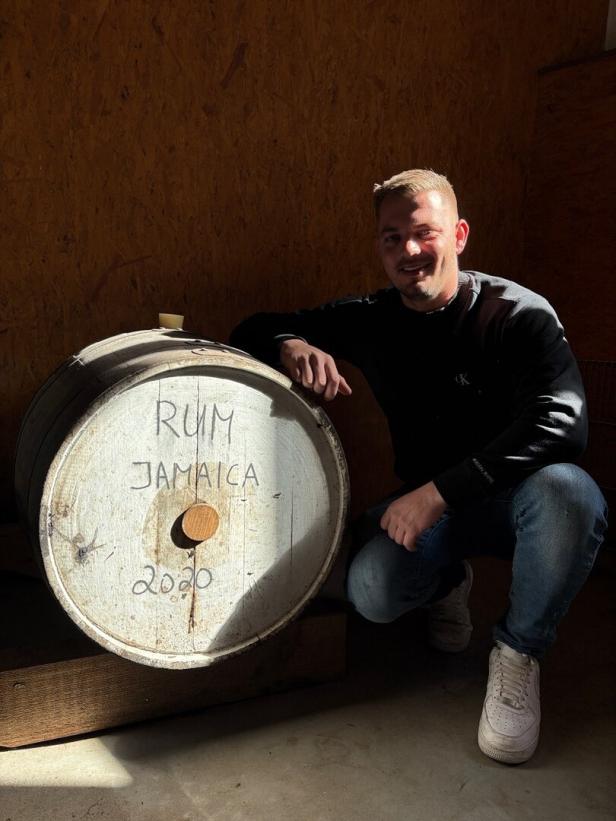 Ein Mann hockt neben einem Holzfass mit der Aufschrift „Rum Jamaica 2020“ in einem Raum mit Holzverkleidung.